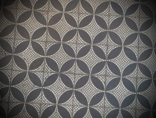 Fotografija geometričnega vzorca na tleh, sestavljenega iz mozaika belih in črnih kamnov na ploščicah. Gre za medseboj povezane štirikotne bele zvezde, obkrožene s črnimi krogi za njimi.