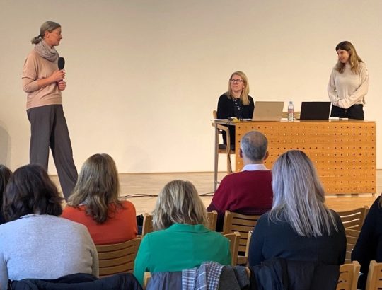 dekorativna slika - konferenca. Na odru sta dve osebi za mizo z računalniki, ena pa stoji in ima v rokah mikrofon. Poslušalci sedijo na lesenih stolih in so vidni v hrbet.