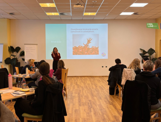 Fotografija usposabljanja za vodje v procesih dezinstitucionalizacije. Pred projekcijo, ki ima naslov "Zmanjševanje birokracije poveča (z)možnosti in ustvarjalnost", stoji predavateljica. Po prostoru so za mizami po skupinah posedeni udeleženci. Na desni zgoraj sta logotipa I feel sLOVEnia in sofinancira EU.