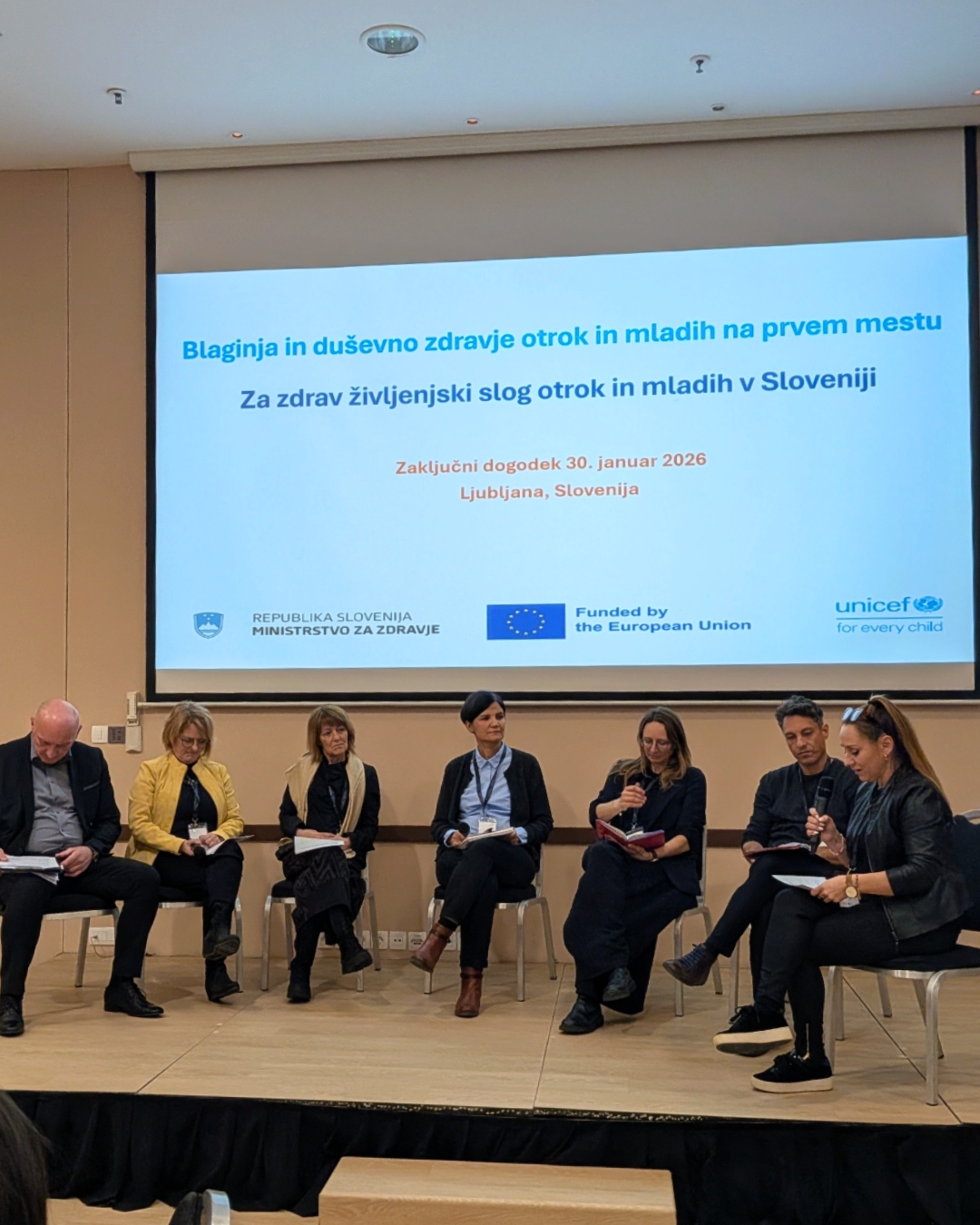 slika iz posveta, udeleženci sedijo na stolih v polkrogu, nad njimi je projekcija predstavitve dogodka